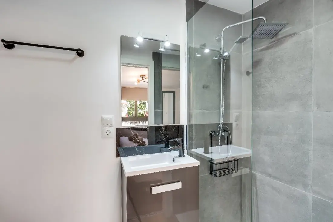 The bathroom in grey tile, with a basin under a long mirror and a glass walk-in shower