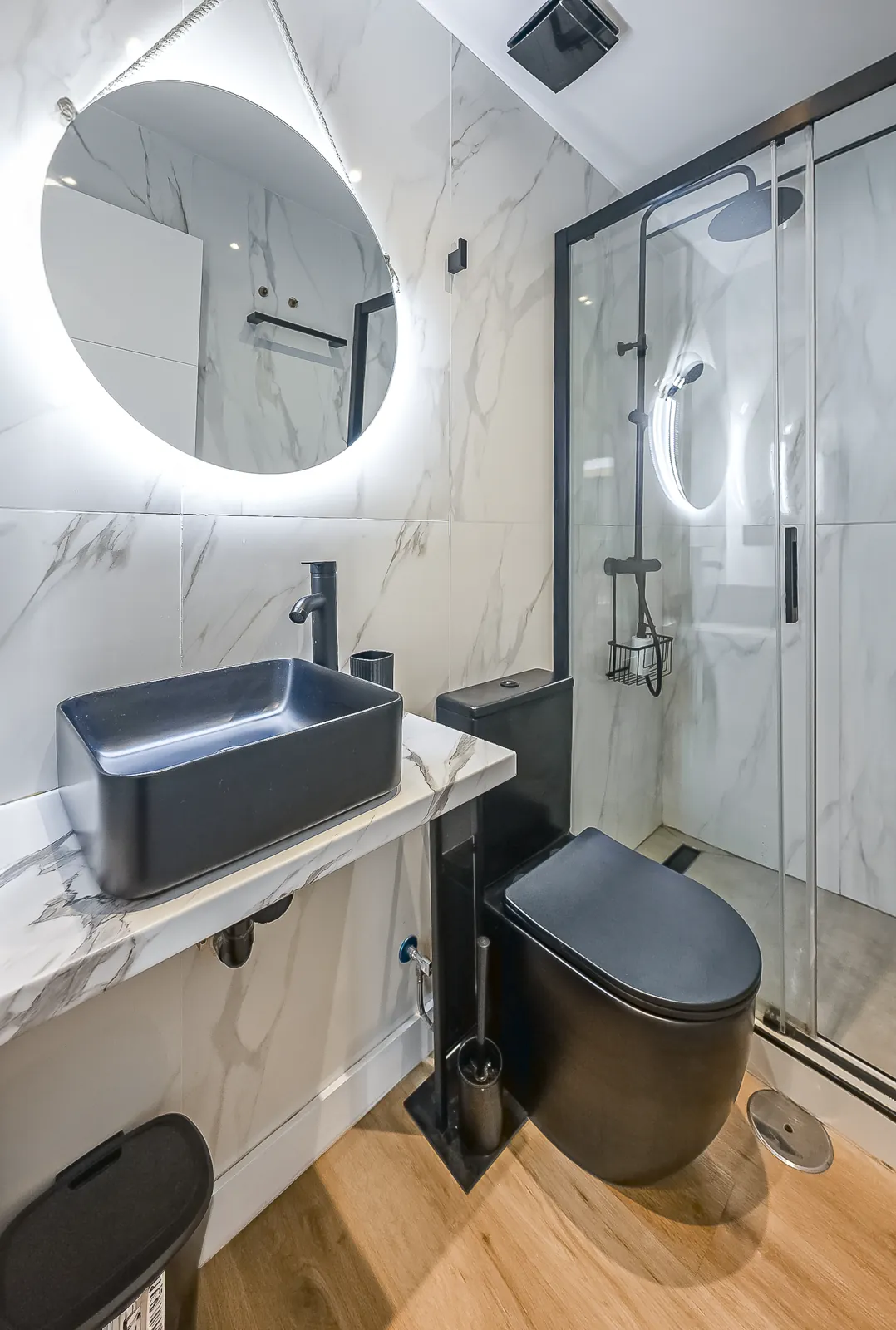 A Signature Suite bathroom: a round lit mirror and a black stone basin on marble, beside the glass shower