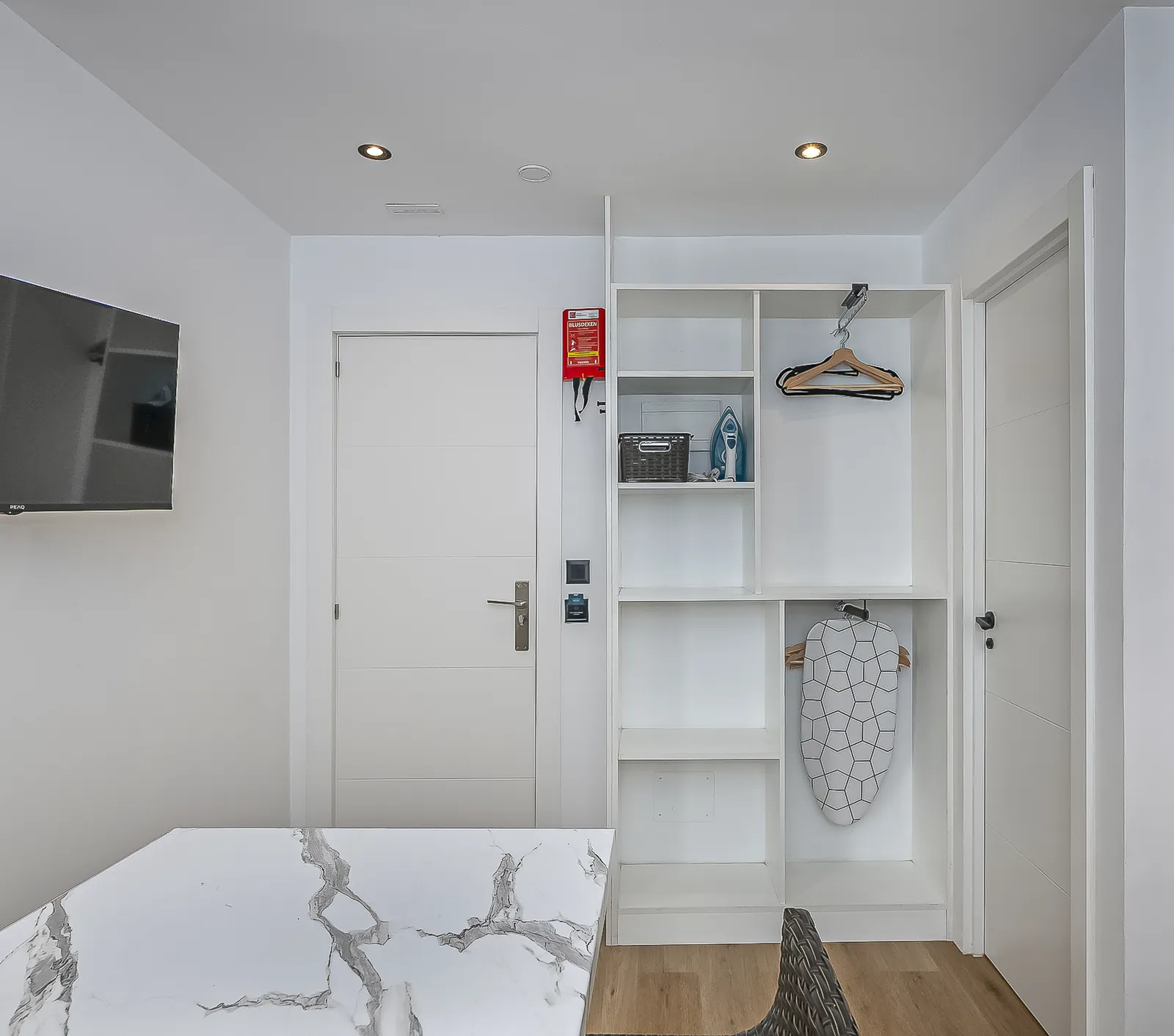 Room 1: open white shelving with hangers, an iron and a safe, beside the door to the bathroom