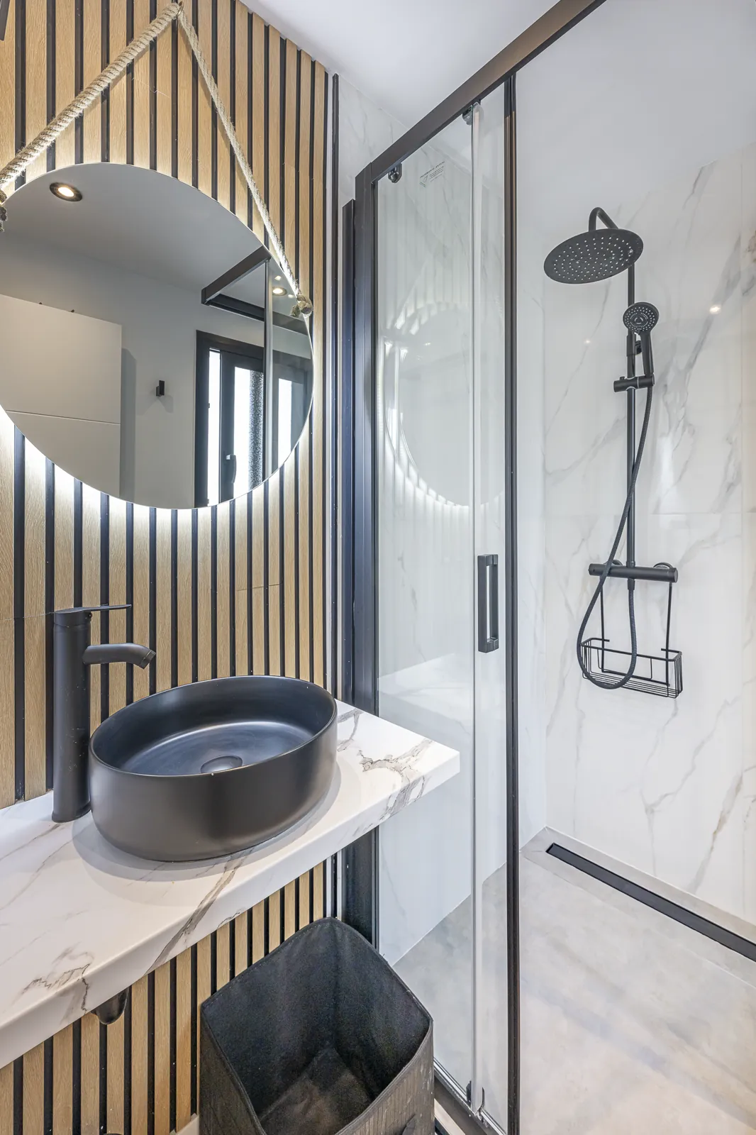 Room 4: the bathroom against oak slats, a round lit mirror over a black stone basin, and the glass shower
